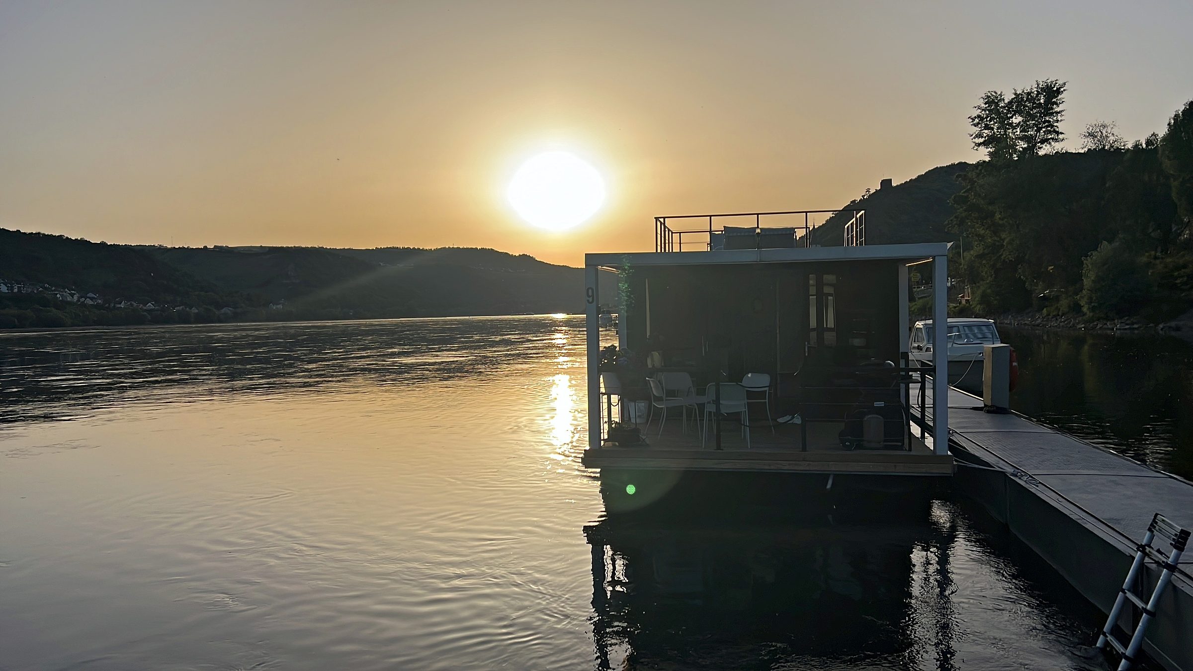 Sonnenuntergang über dem Rhein; das Hausboot liegt im Gegenlicht am Steg, die Sonne spiegelt sich als Bahn auf dem Wasser.