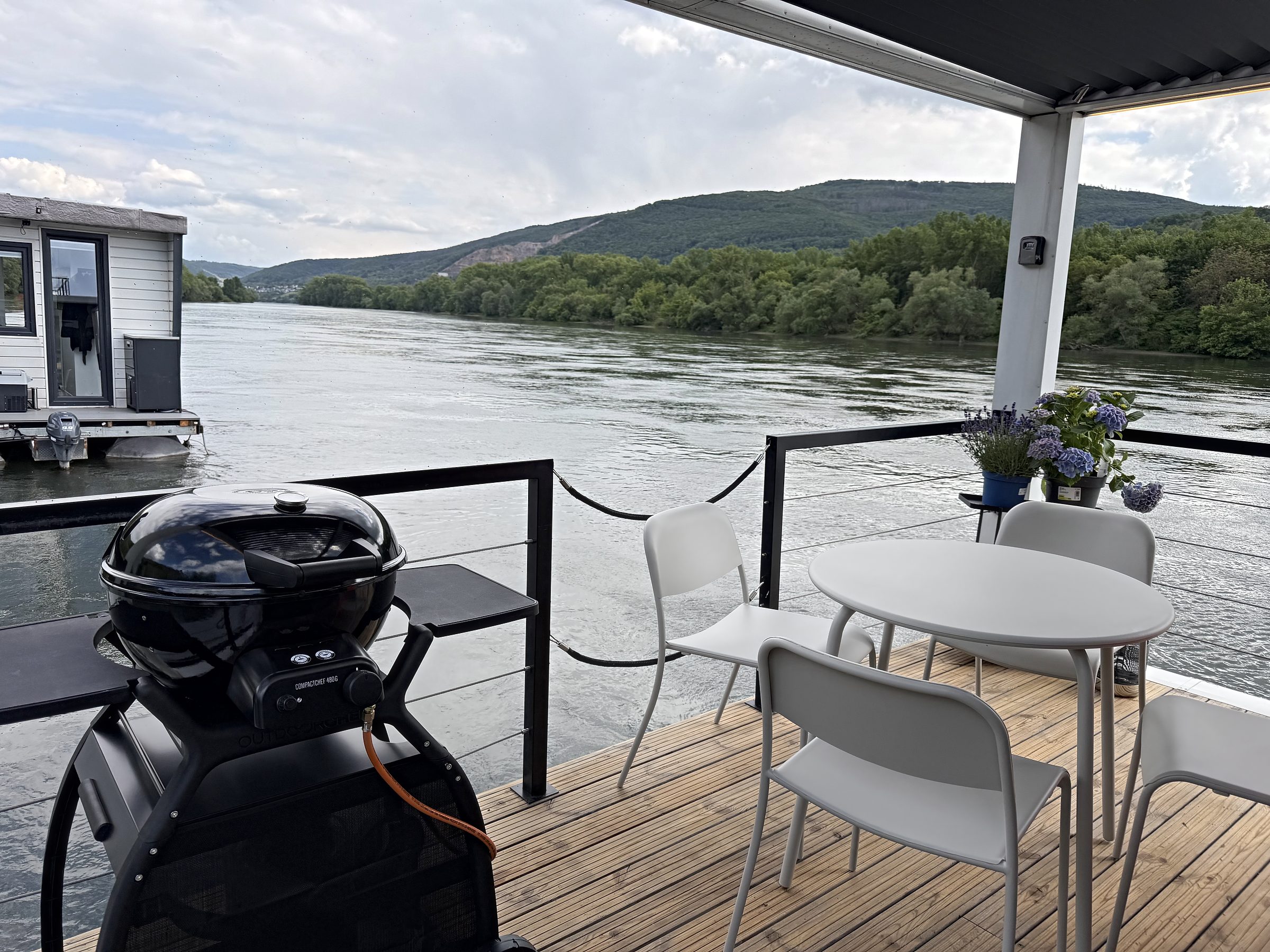 Die Dachterrasse von Modern 1 mit Gasgrill, Tisch, Stühlen und Hortensien, dahinter der Rhein und die bewaldeten Hänge.