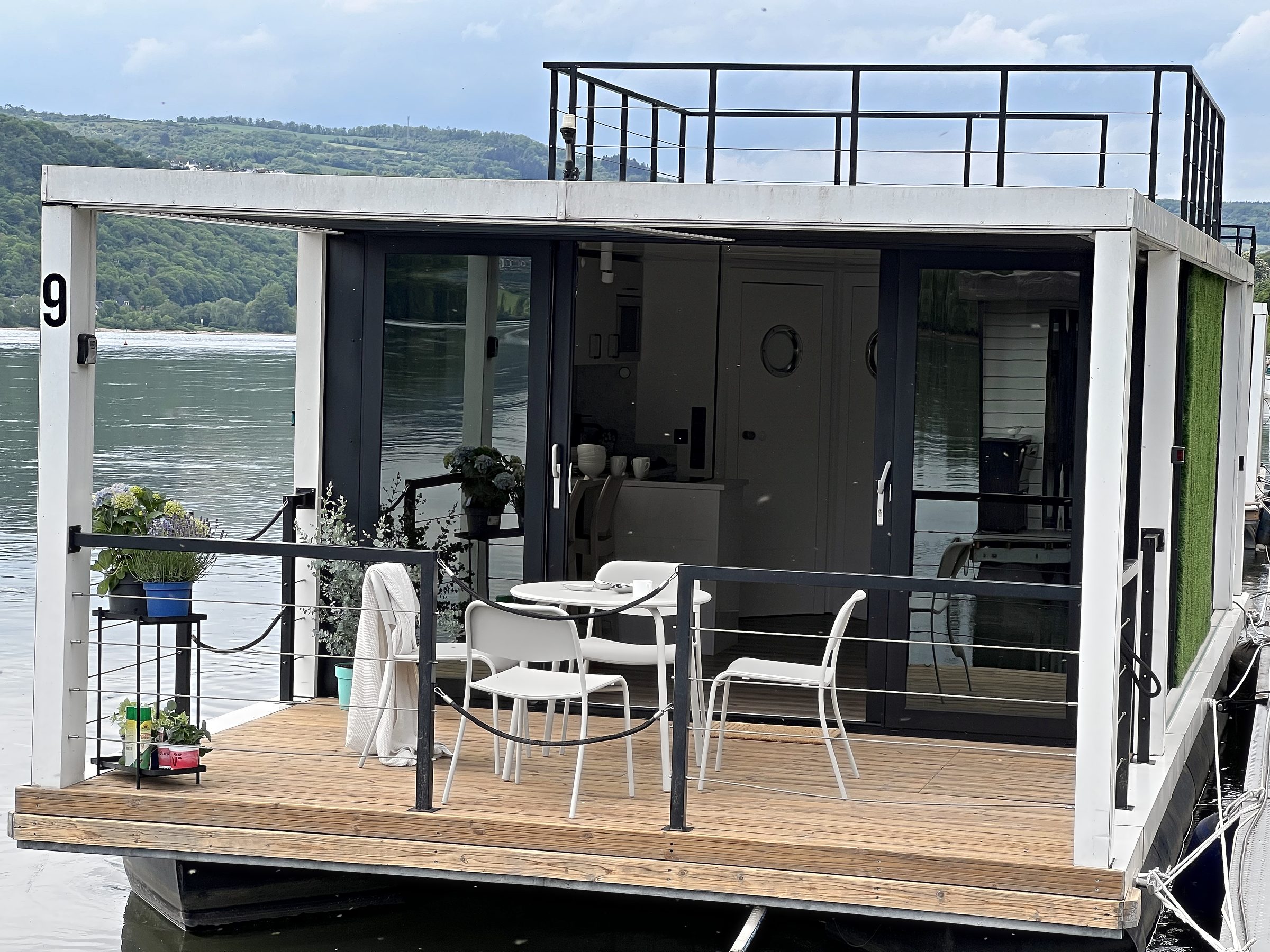 Das Hausboot Modern 2 von außen: weiße Fassade, Holzterrasse mit Tisch und Stühlen, darüber die begehbare Dachterrasse.