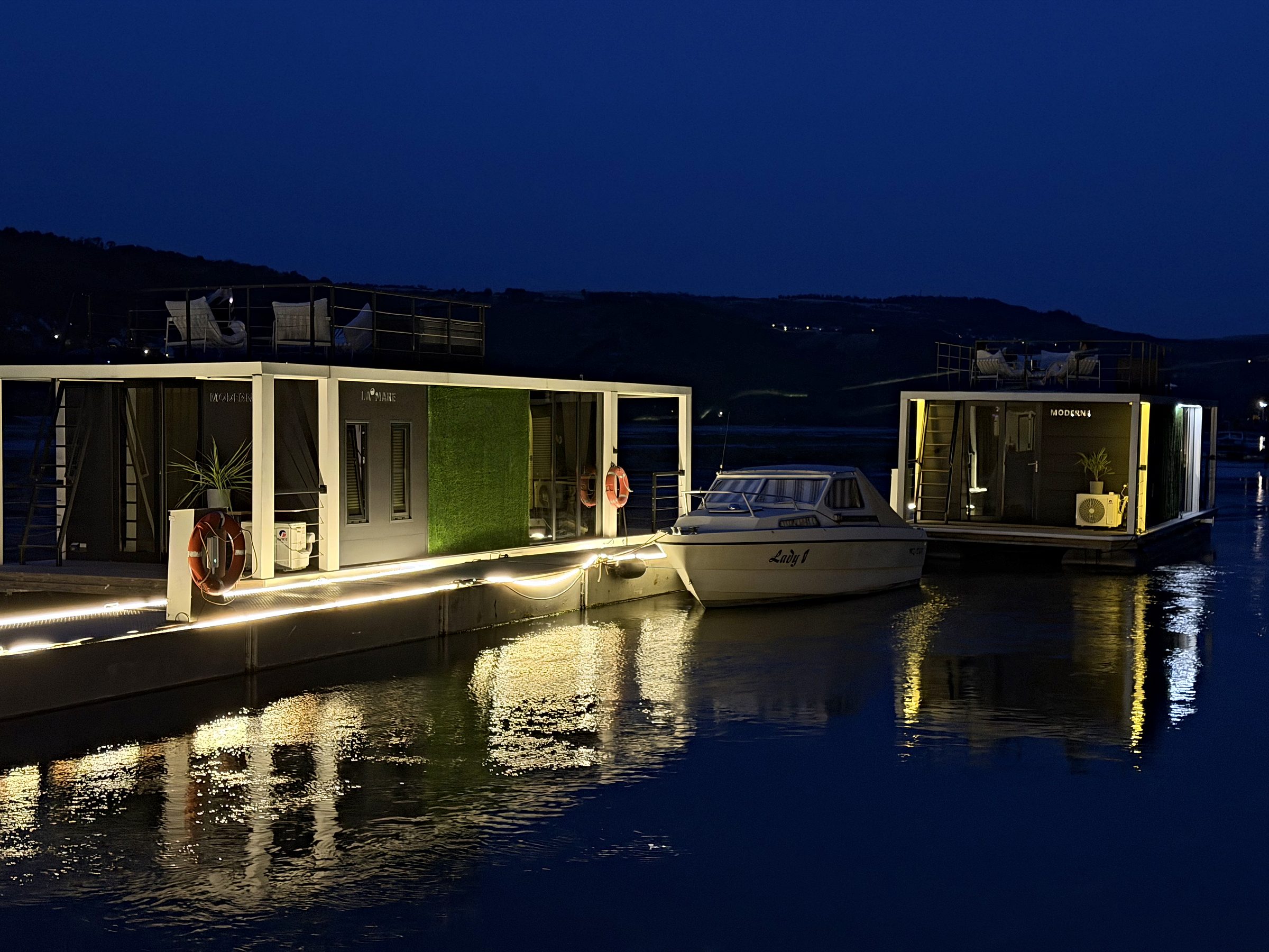 Beide Hausboote der Burgflotte, Modern 1 und Modern 2, liegen in der blauen Stunde beleuchtet am Steg der Marina Rheinlicht; das warme Licht spiegelt sich im ruhigen Rhein.