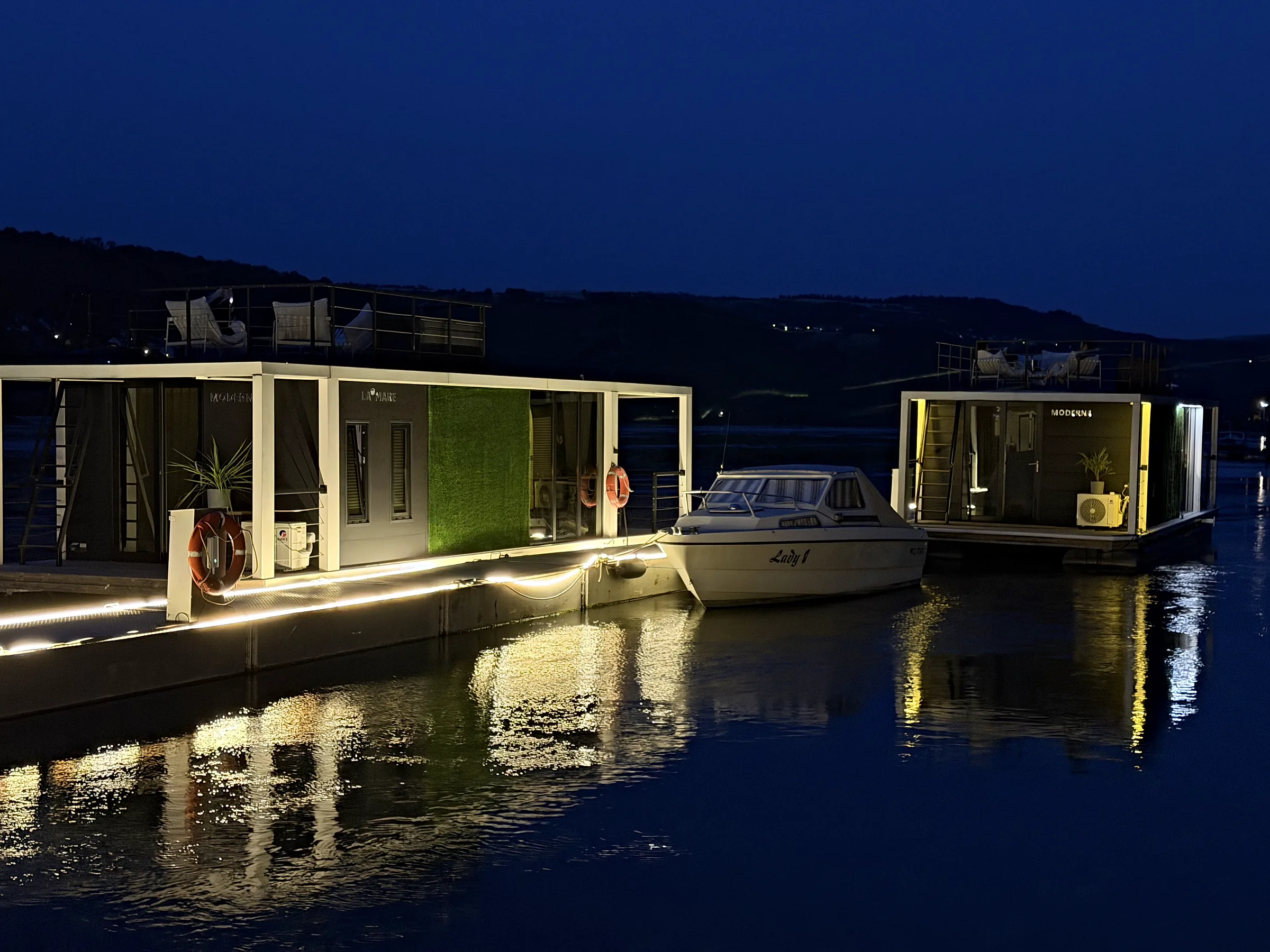 Beide Hausboote der Burgflotte, Modern 1 und Modern 2, liegen in der blauen Stunde beleuchtet am Steg der Marina Rheinlicht; das warme Licht spiegelt sich im ruhigen Rhein.