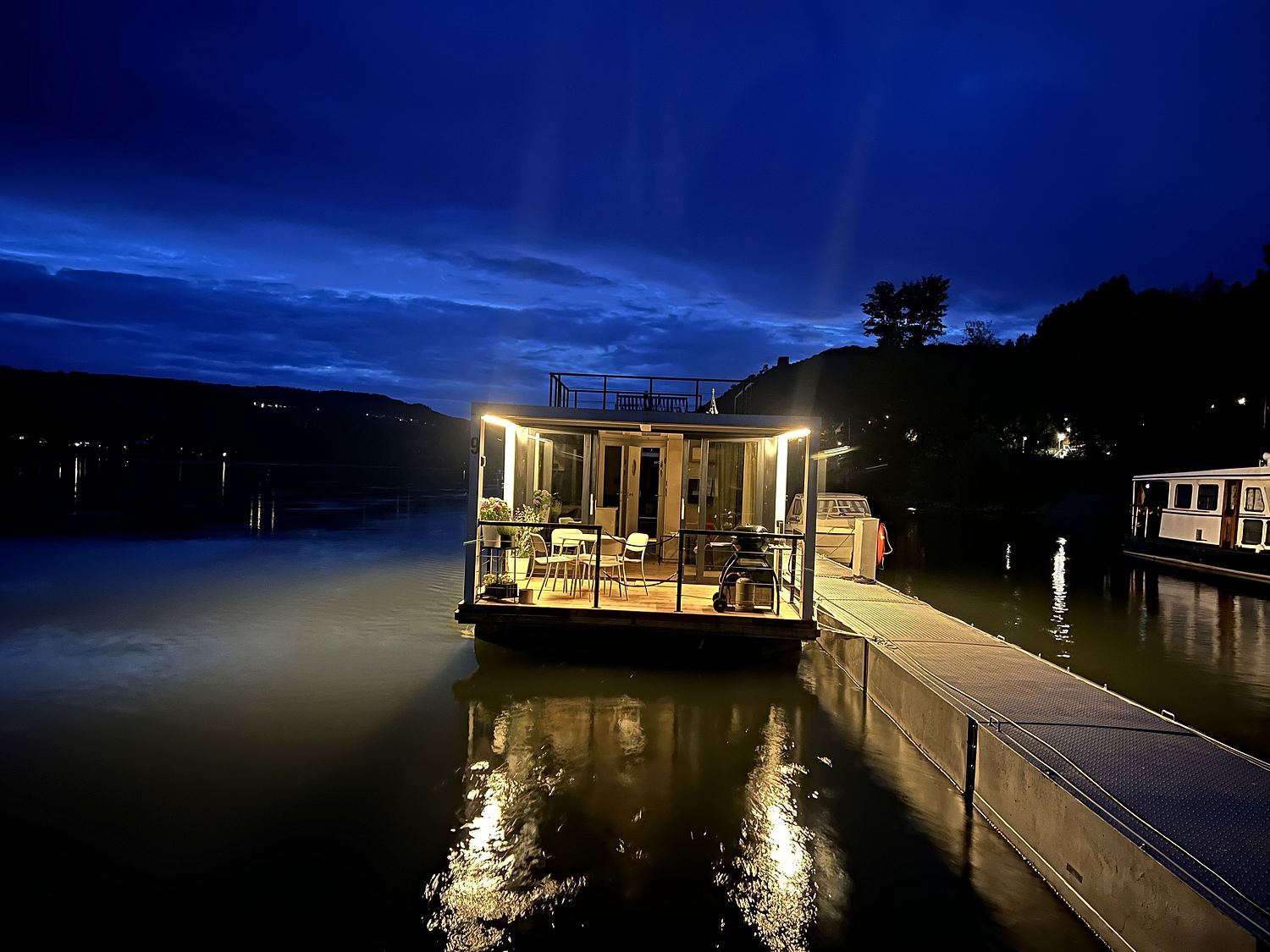 Ein Hausboot bei Nacht, warmes Licht fällt aus den Panoramafenstern auf das Wasser.